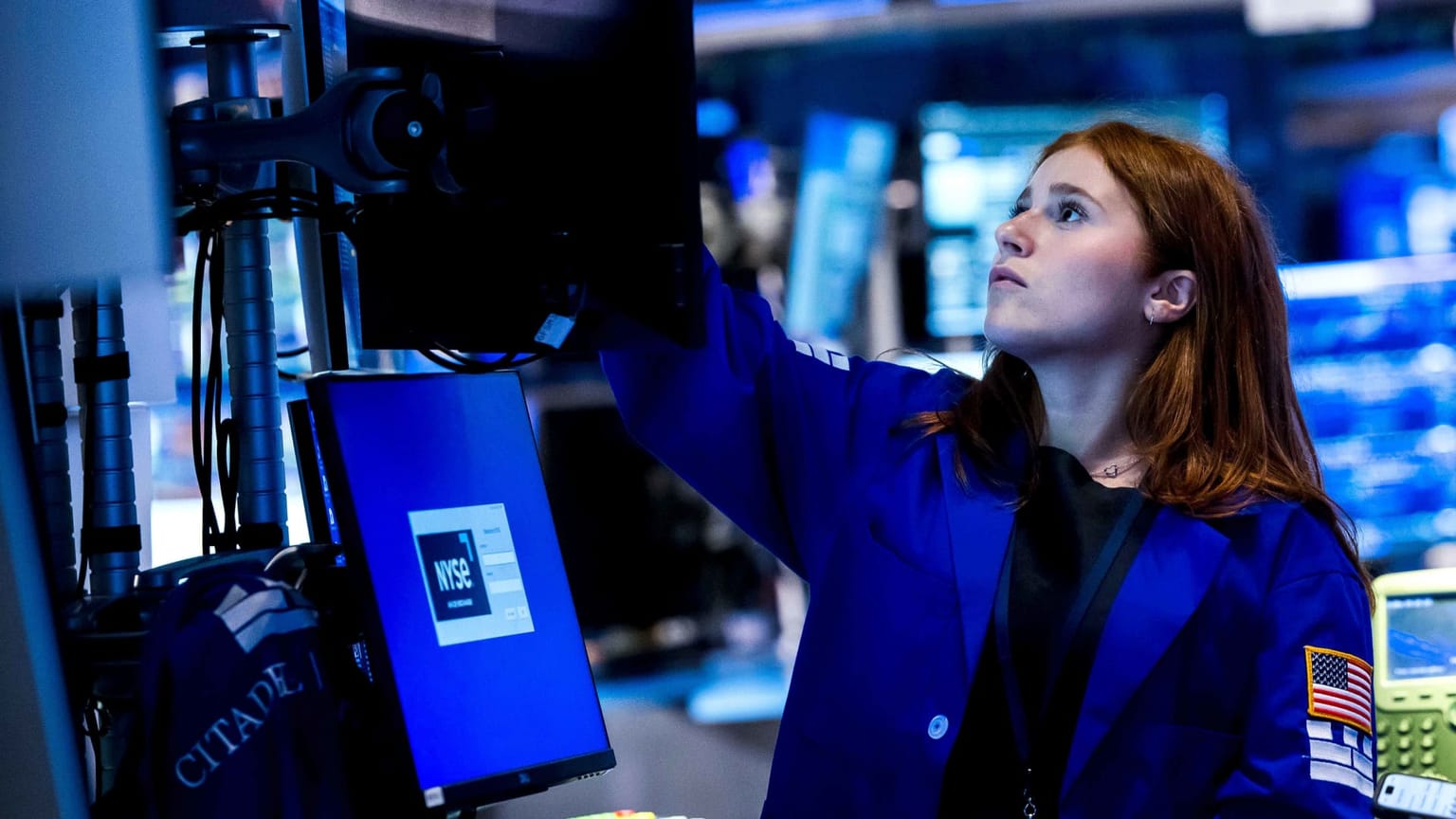 A New York Stock Exchange floor trader in a blue jacket reaches up to adjust a monitor on June 26 2026, illustrating the ThriveInMarkets evening review after US stocks tread water despite a deepening global technology selloff, with the S&P 500 closing essentially flat at 7,354.02, the Nasdaq Composite slipping 0.24 percent to 25,297.62 for a fifth straight loss and the Dow easing 0.09 percent to 51,876.11, as a New York Times report that OpenAI may delay its public listing into next year pressured chip stocks alongside Apple and Microsoft price hikes, while Bitcoin probed its lowest level since September 2024 near 58,189 dollars before recovering to about 59,632, Ethereum eased to about 1,569 dollars, gold reclaimed ground near 4,068 dollars and WTI crude slumped about 3.5 percent toward 69.40