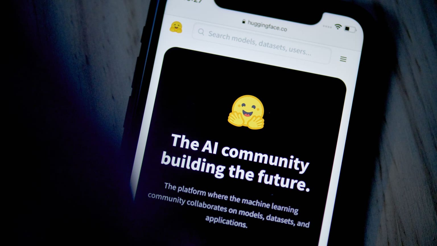 A smartphone showing the Hugging Face website with the yellow hugging-face emoji and the headline The AI community building the future, used as the ThriveInMarkets evening-review inline image for Thursday August 27 2026, when The Information reported Nvidia had agreed to buy the open-source model hub for 12.9 billion dollars, a deal neither company confirmed, as Nvidia shares closed about 9 percent higher after Wednesday night's 70 percent fiscal 2028 growth guide