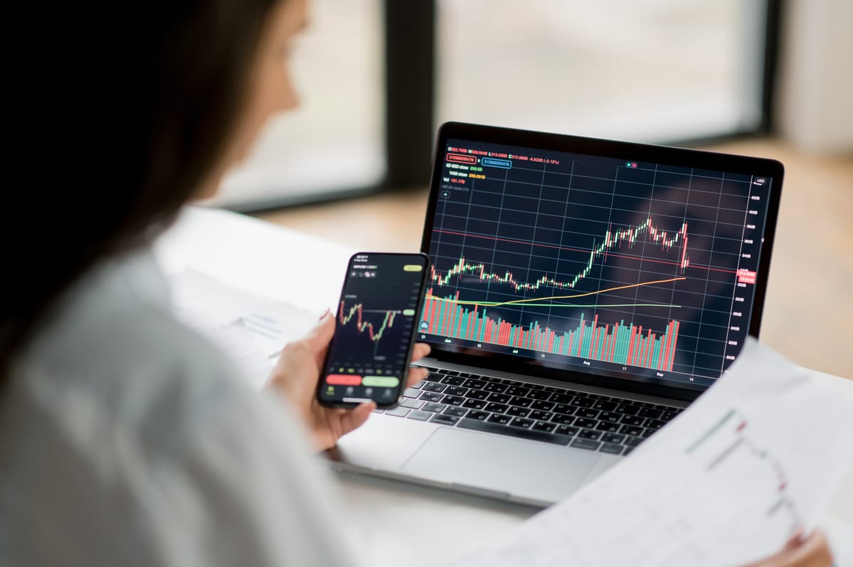 A trader studies candlestick charts on a laptop and a phone, used as the ThriveInMarkets evening-review hero for Monday August 31 2026, when the S&P 500 closed 7,677, down 0.45 percent, as West Texas Intermediate held near 85.60 dollars after Sunday US strikes on Iranian launchers at Larak Island and Tesla jumped about 5 percent, leaving August still in the green