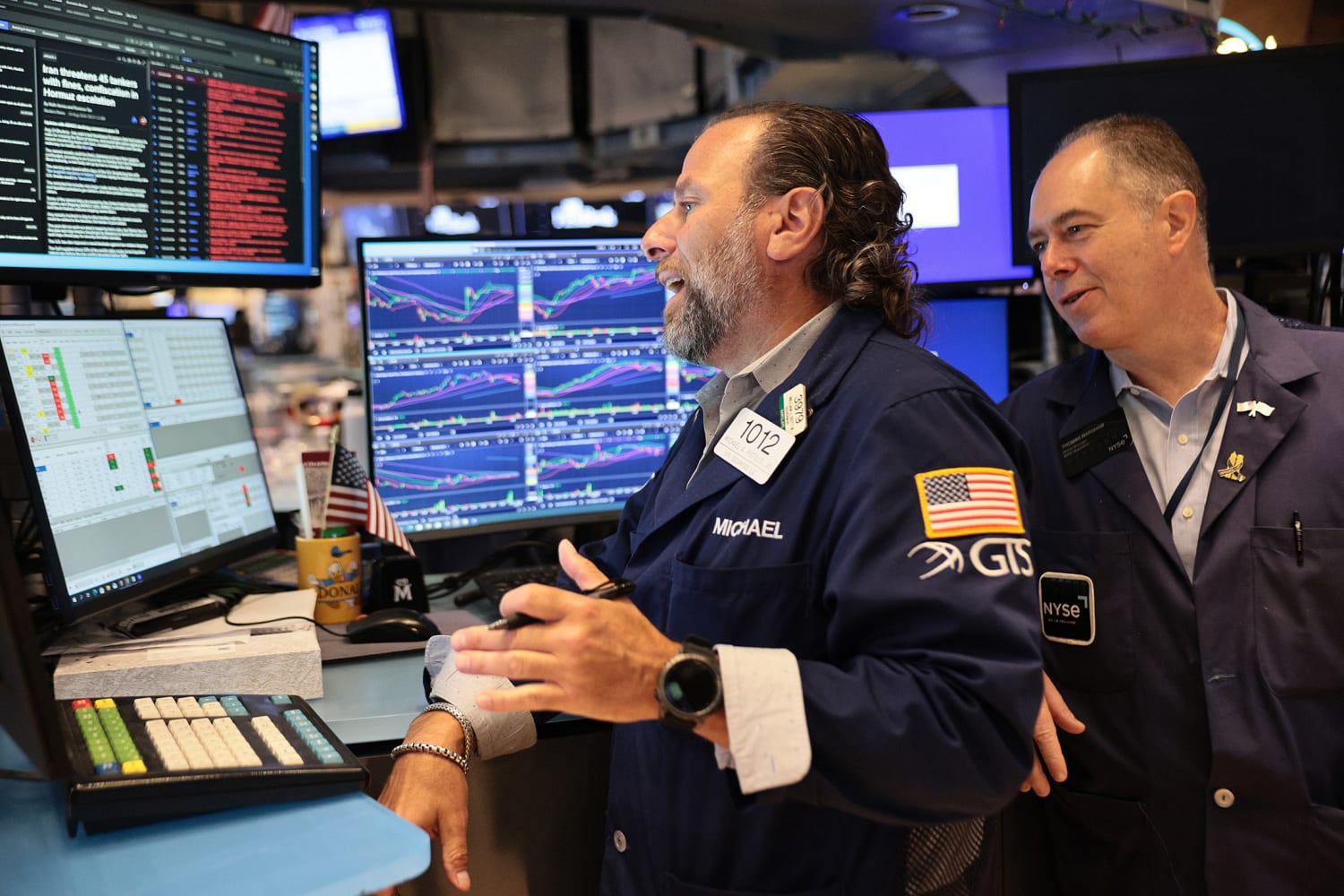 NYSE floor traders including Michael, badge 1012 of GTS, stand at a bank of quote screens showing charts and an Iran-Hormuz headline on 21 September 2026, used as the ThriveInMarkets evening-review hero when the Nasdaq Composite closed 27,122.09 at a record, Advanced Micro Devices reached a 1 trillion dollar market value, the S&P 500 closed 7,764.70, and West Texas Intermediate last printed about 95.80 dollars