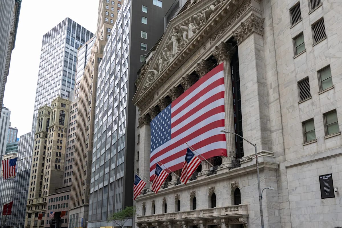 The New York Stock Exchange facade on Broad Street with a large American flag across the columns, used as the ThriveInMarkets evening-review hero for Thursday October 15 2026, when the S&P 500 closed 7,744.06 up 0.33 percent after September PPI jumped 1.3 percent month on month, the largest gain since March 2022