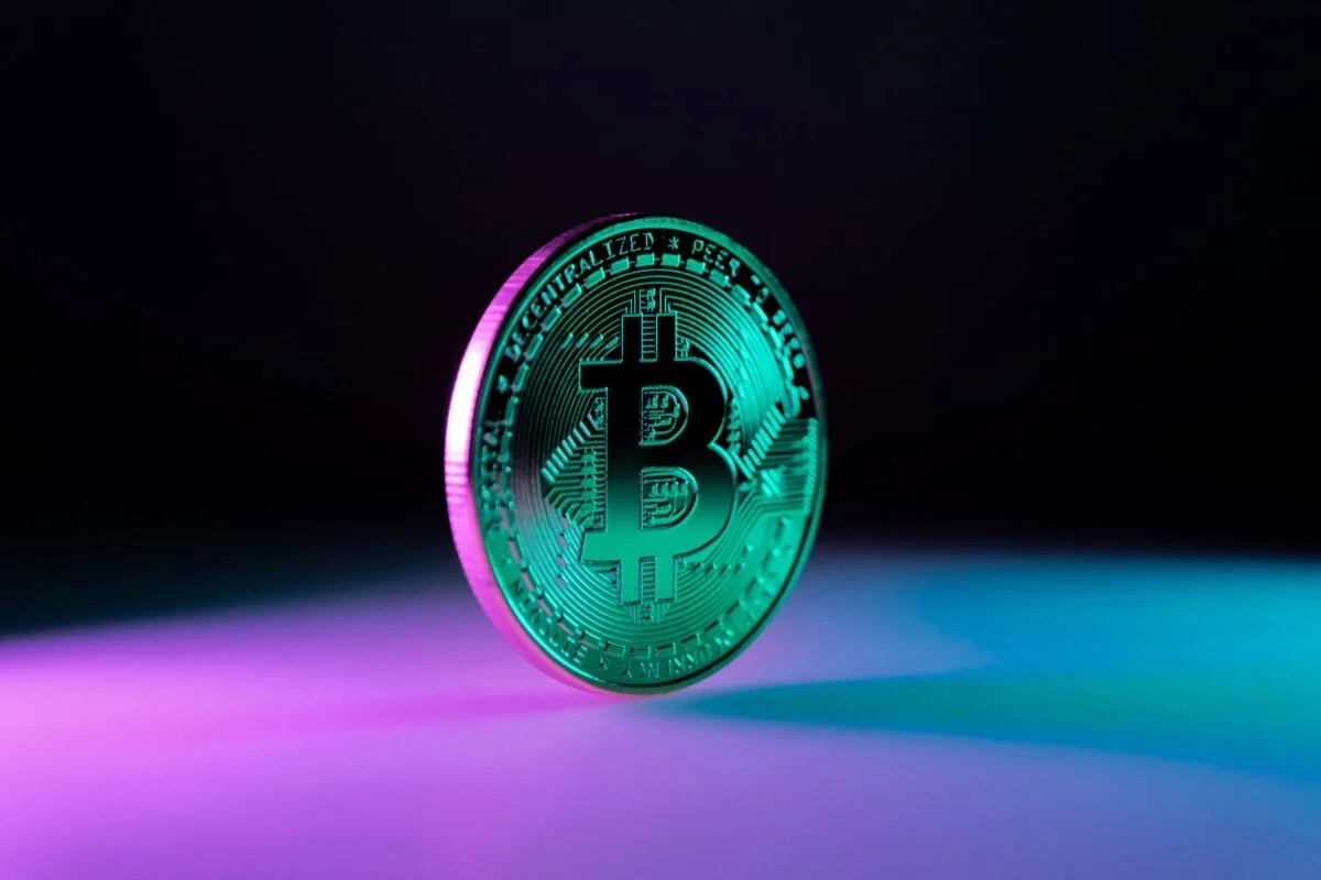A polished metallic Bitcoin coin stands upright on a dark surface lit by neon teal and magenta light, illustrating the June 19 2026 ThriveInMarkets morning analysis as Bitcoin slipped below 63,000 dollars to about 62,567 dollars, down roughly 2.8 percent over 24 hours, with more than a billion dollars in leveraged longs liquidated as a hawkish Federal Reserve dot plot offset the US-Iran peace deal, Ethereum traded near 1,696 dollars, spot gold eased to about 4,157 dollars after recovering above 4,300 on Thursday and WTI crude hovered near 75 dollars close to three-month lows, all while US stock and bond markets stayed closed for the Juneteenth holiday and reopen Monday June 22