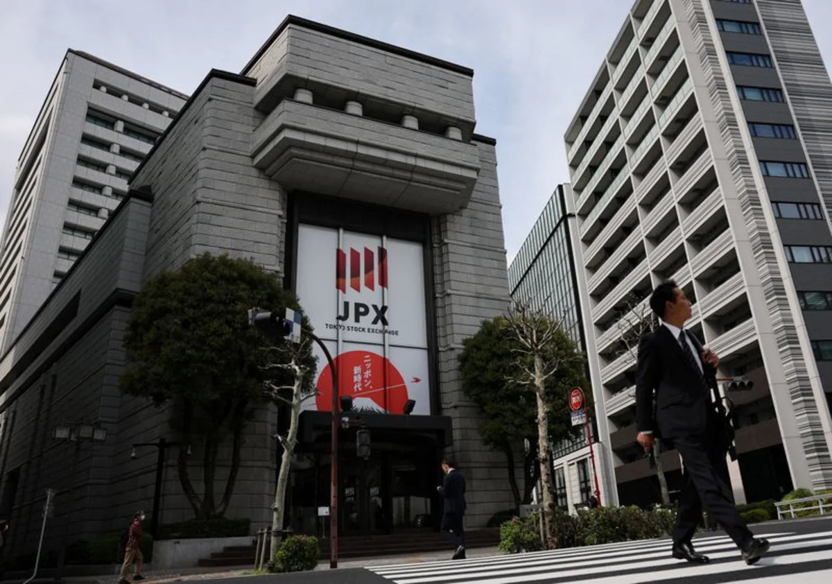 The Tokyo Stock Exchange building with JPX branding as a pedestrian in a dark suit crosses the street, marking Friday August 14 2026 as Asian stocks rose and US index futures held little changed after the S&P 500 closed Thursday at a record 7,798.99 on a flat July Producer Price Index, with July retail sales and the preliminary August University of Michigan consumer sentiment survey still to come